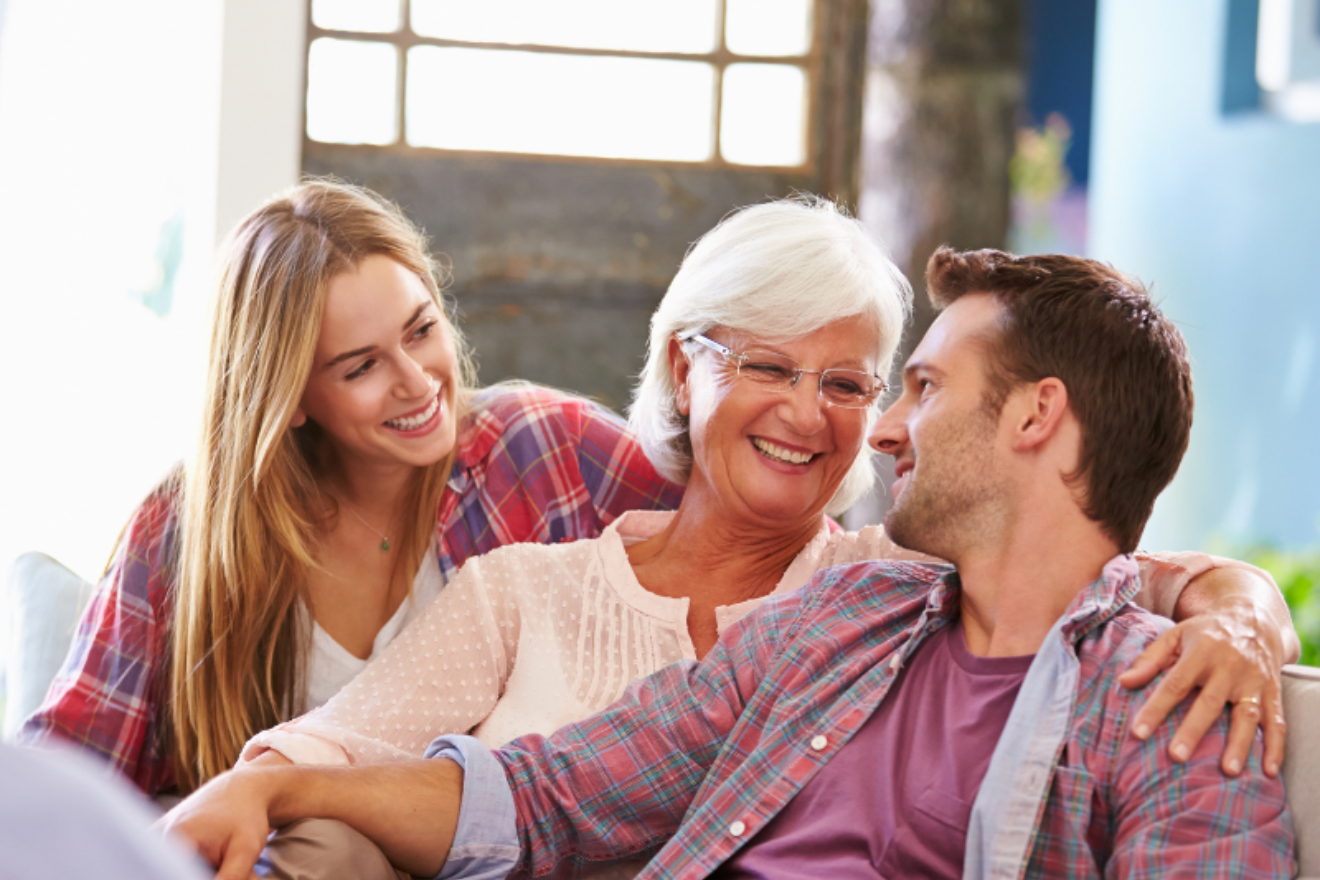 A photograph of an elderly woman smiling on a couch, seated between her two adult children and with her arm around her son.