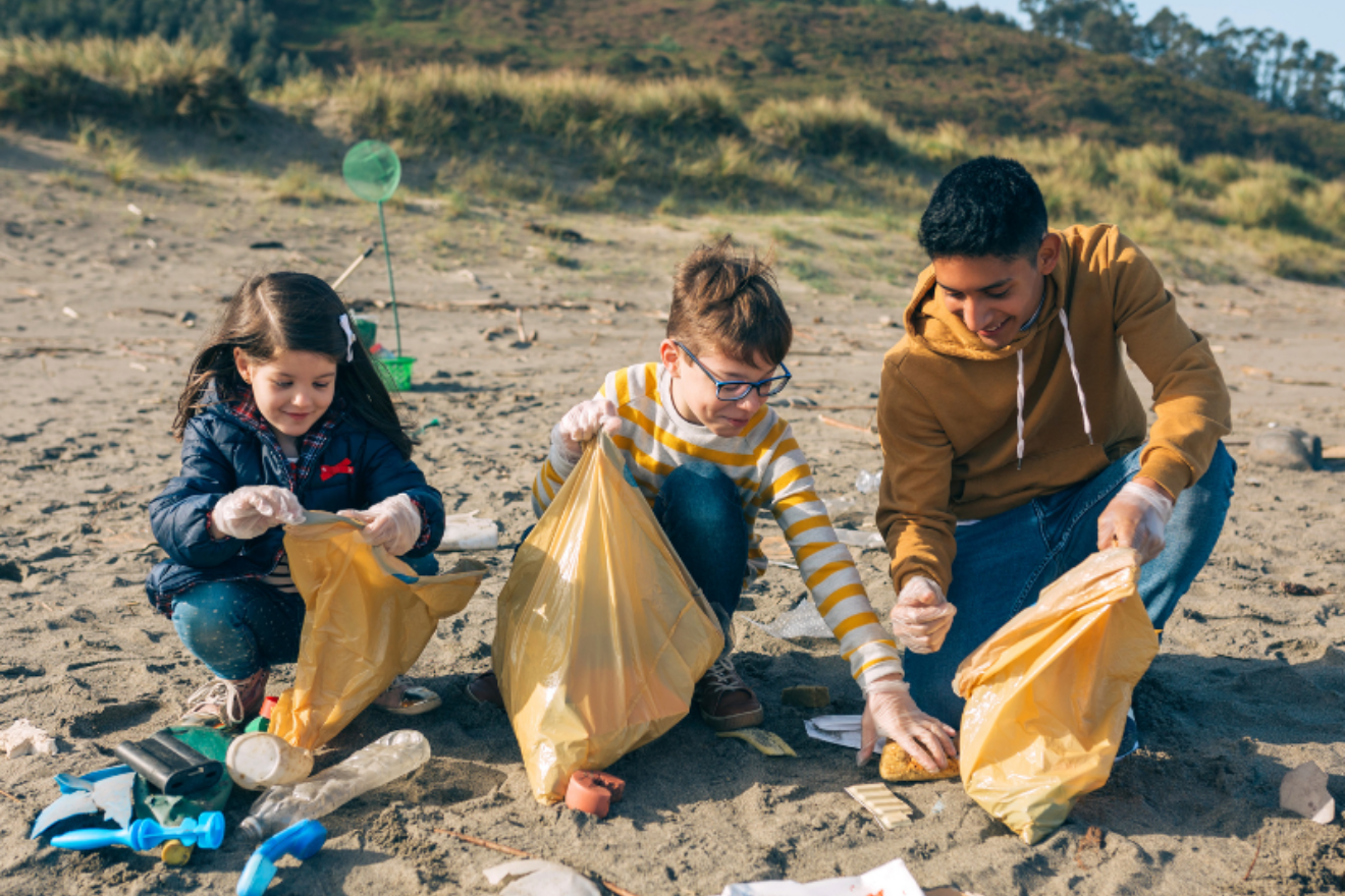 A photograph of a young man and two small children picking litter up off a beach and placing it into bags.