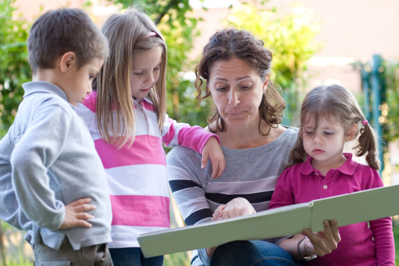A photograph of a woman holding a picture book or photo album in front of her three young children. All have disheartened expressions on their faces.