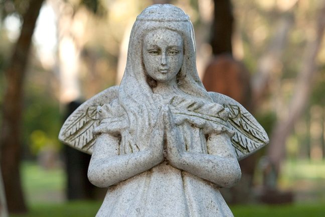 A photograph of a stone monument, featuring an angel with her head bowed and hands folded in prayer.