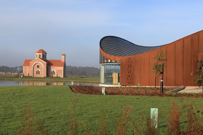 A photograph of the Sanctuary building at Bunurong Memorial Park, surrounded by sweeping lawns. The Saint Lazarus Greek Orthodox Church is visible in the background. A blue sky completes the serene setting.