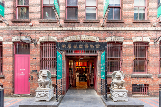 A photograph of the facade of the Chinese museum building, with the open front door at the centre. The doorway is flanked by two ornate statues.