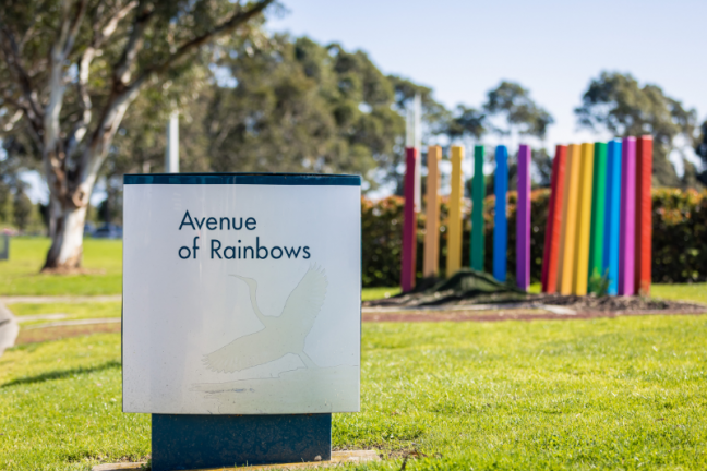A photograph of the entry signage at Avenue of Rainbows. The sign sits on a bright, green lawn with a decorative rainbow sculpture in the background. Gum trees stand proudly behind the sculpture, against a blue sky.