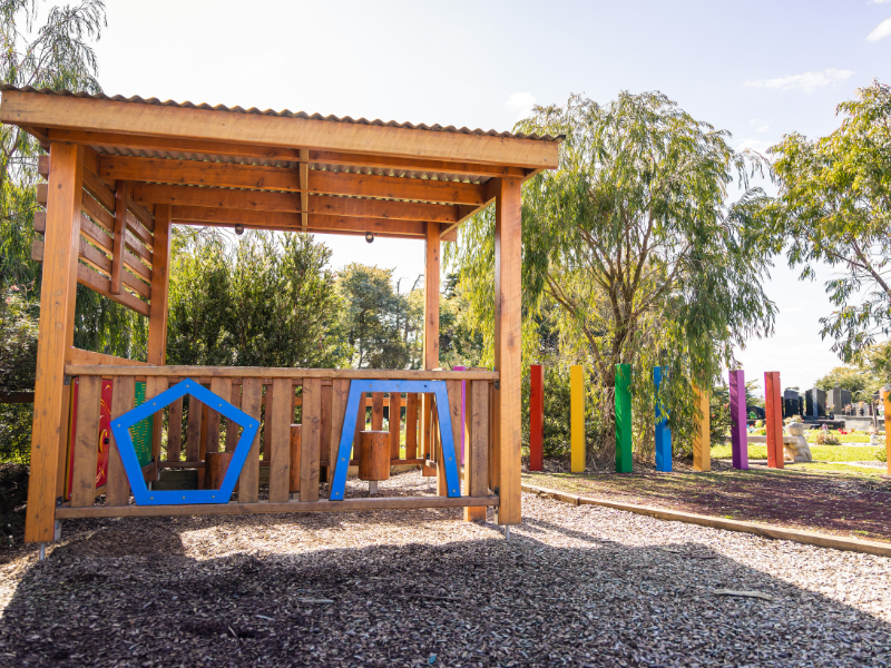 A photograph of the cubby house at Avenue of Rainbows. The cubby is built from golden-toned timber with colourful accents. The sun beams down and the sky is blue.