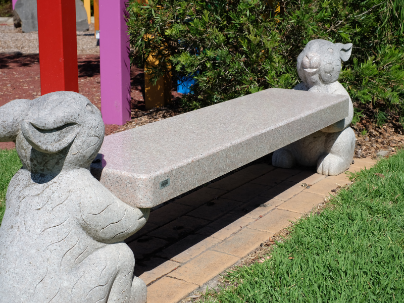 A photograph of a low, grey stone bench at Avenue of Rainbows. Whimsical rabbit sculptures lie on opposite ends of the bench.
