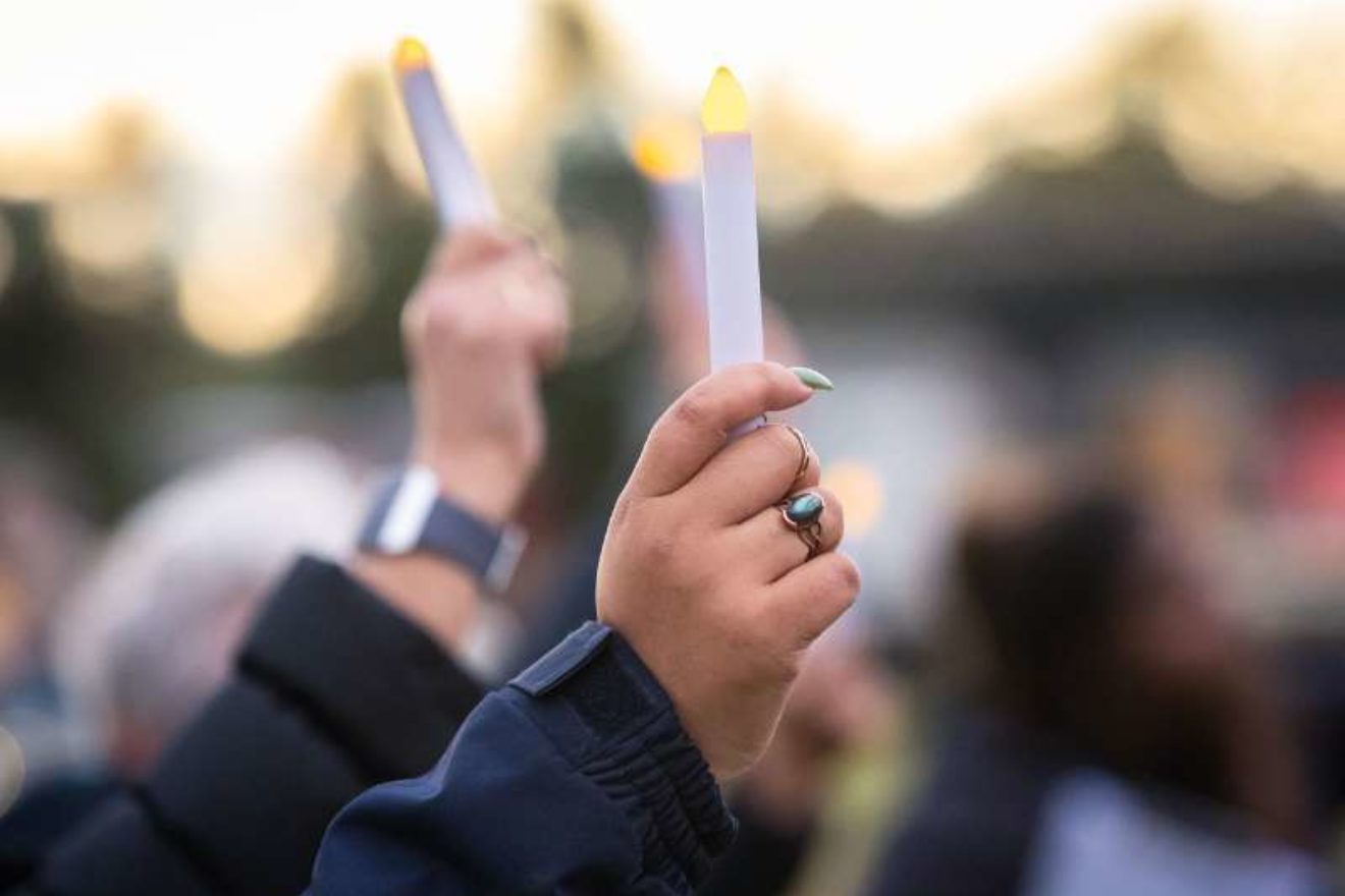 A close-up photograph of two hands holding LED candles at an outdoor remembrance event.