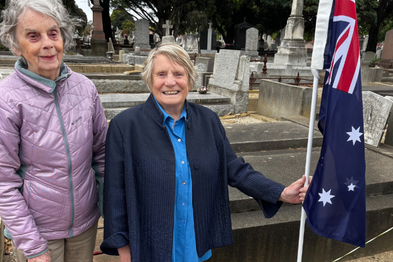 Two people facing forward standing in front of graves at Brighton General Cemetery. The person on the right holds a large Australian flag in their hand.
