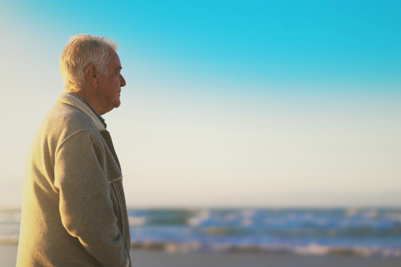 A photograph of a middle-aged man wearing a beige coat and gazing out over a calm ocean. He has a neutral expression.