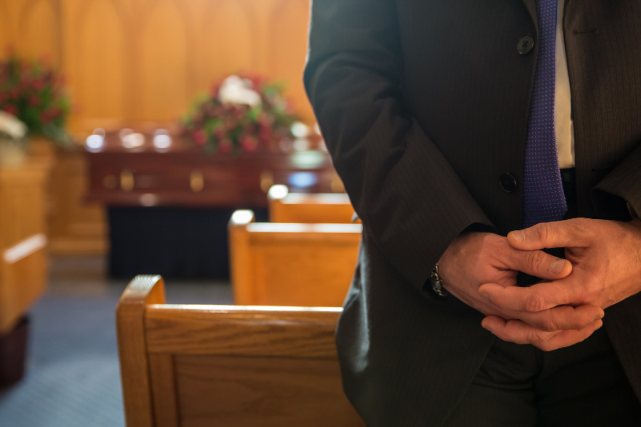 A funeral director standing in a chapel with hands clasped, photographed from the shoulders down