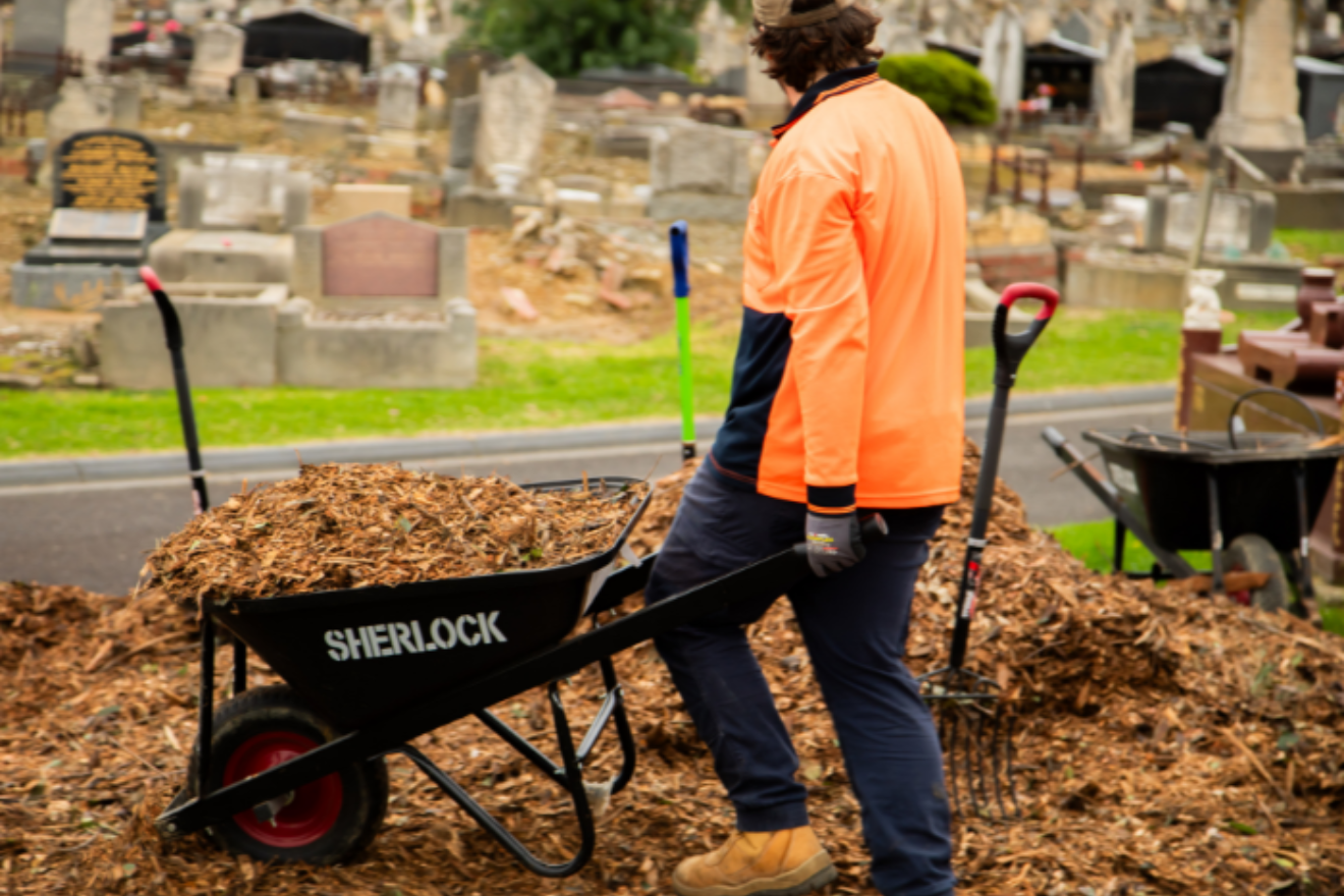 A person in a hi-vis orange jacket stands with their back to the camera. They are holding the handles of a wheelbarrow of mulch, in front of a large pile of mulch. Gravestones can be seen in the background.