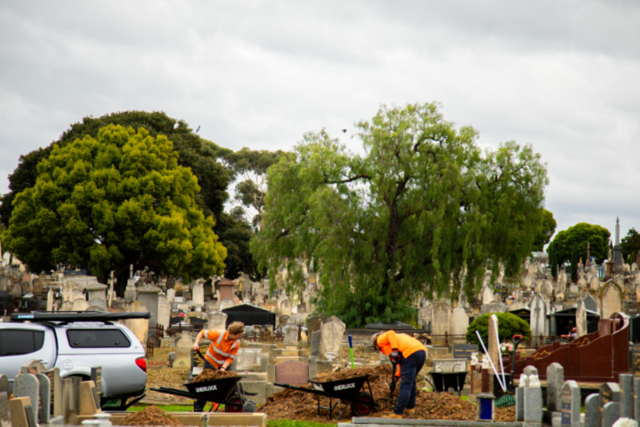 Two people in hi-vis orange jackets bend down as they shovel from a large pile of mulch into two waiting wheelbarrows. In the background can be seen headstones, large leafy green trees and a cloudy overcast sky.