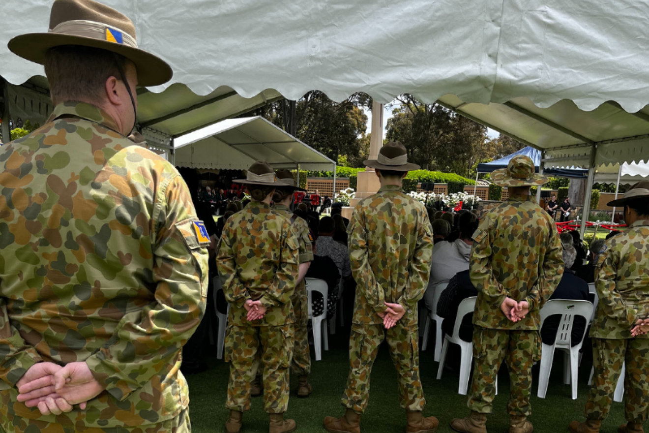 Servicepeople dressed in army fatigues stand at the back of the event marque observing the ceremony with their backs to the camera.
