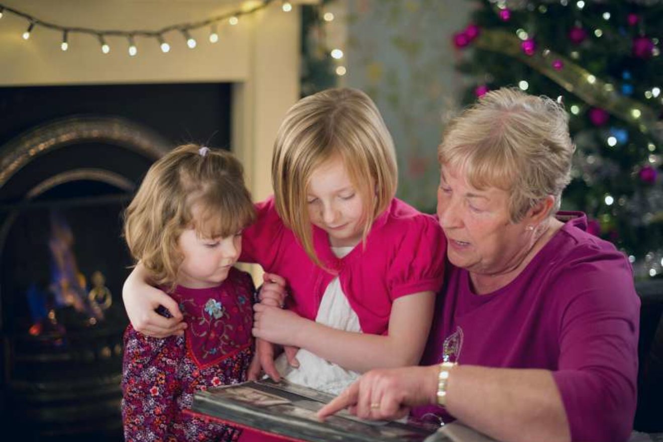 A photograph of a grandmother and two young granddaughters looking at a photo album, with a Christmas tree in the background.