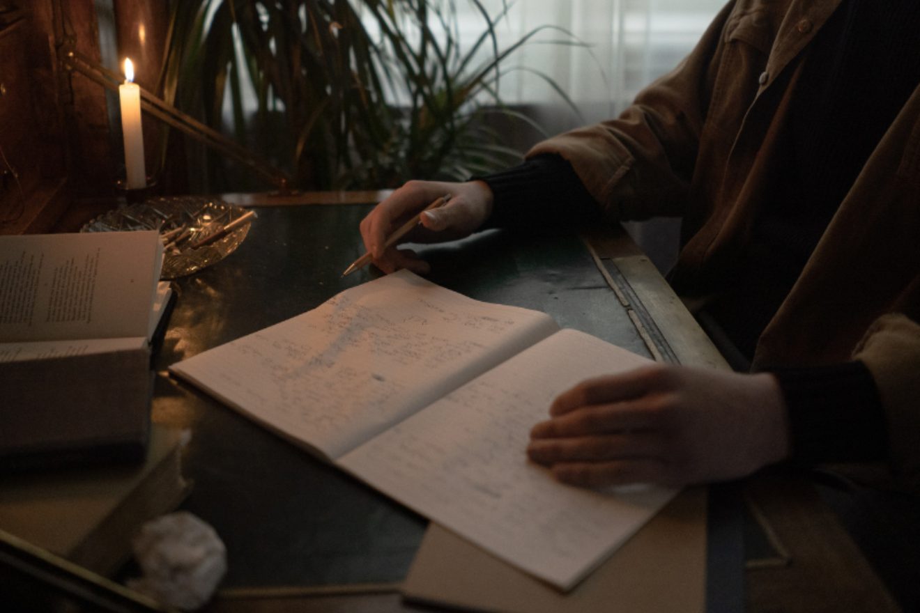 A photograph of a person's hands as they write in a notebook by candlelight.