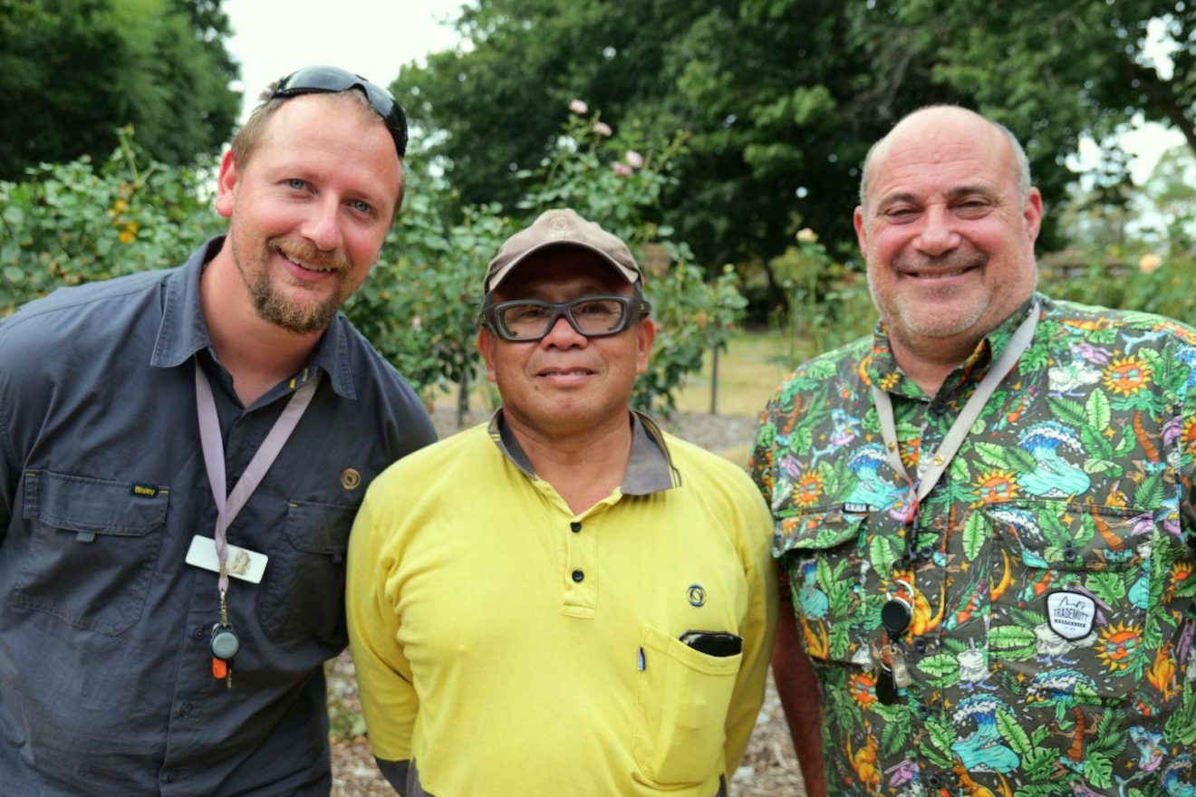 Three men stand together in front of rose bushes.