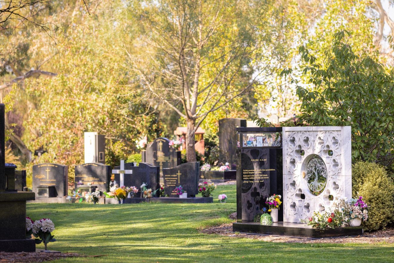 Wide shot of floral arrangements and memorabilia on lawn graves under trees at Springvale Botanical Cemetery.