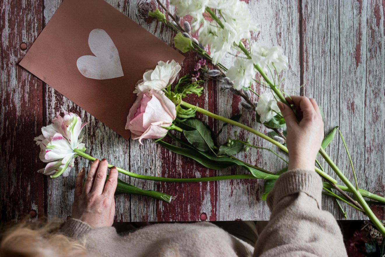 A view from above of a woman preparing roses for a bouquet, with a handmade Valentine's Day greeting card beneath her on the bench.